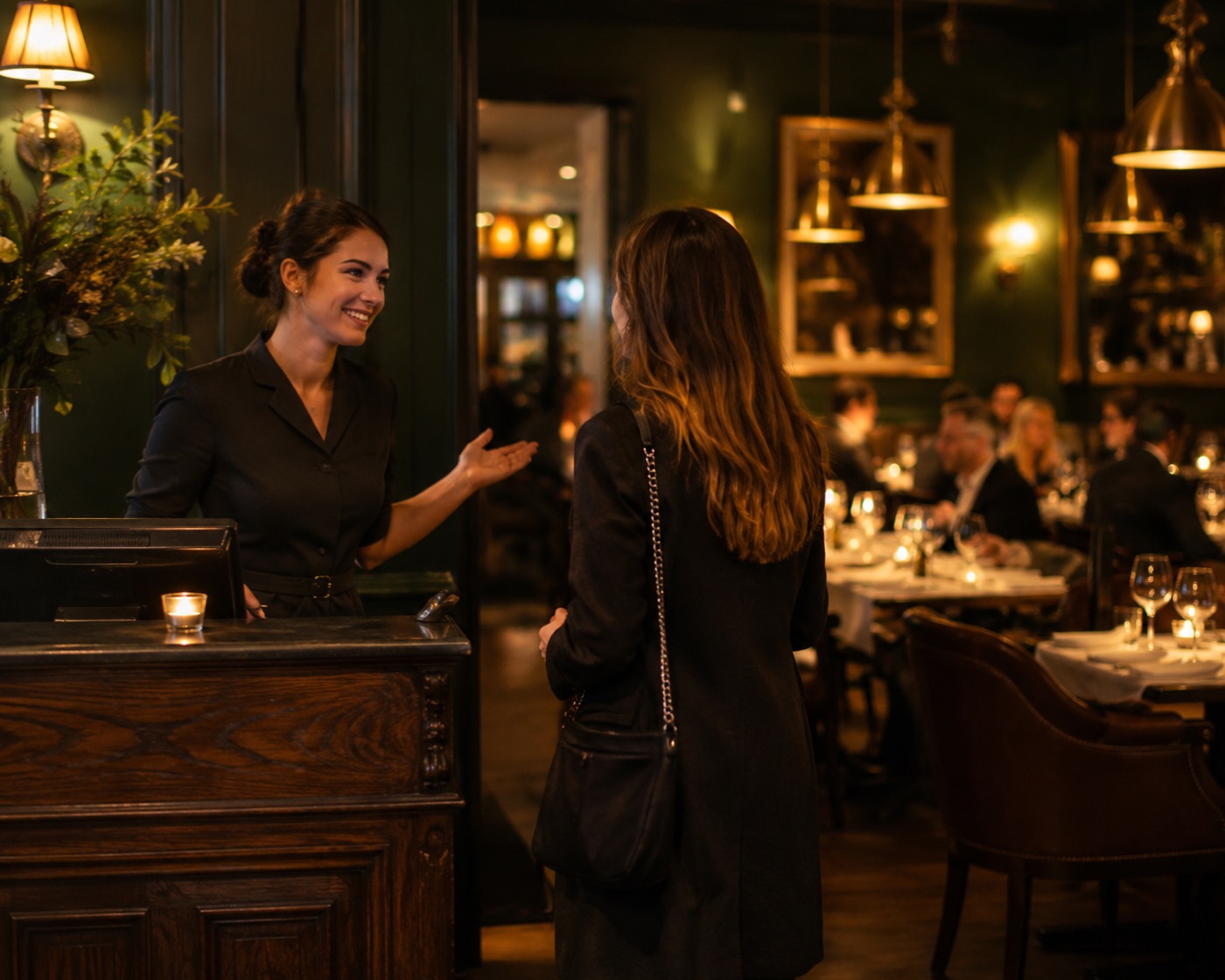 A restaurant host warmly greeting a returning guest at the host stand of a candlelit dining room